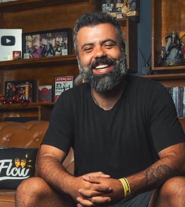 Igor 3k veste OQVestir - Manual do Guarda-Roupa Masculino Igor 3k, apresentador do Flow Podcast, sorrindo e sentado em um sofá de couro marrom, vestindo camiseta preta básica para a campanha Manual do Guarda-Roupa Masculino da OQVestir.