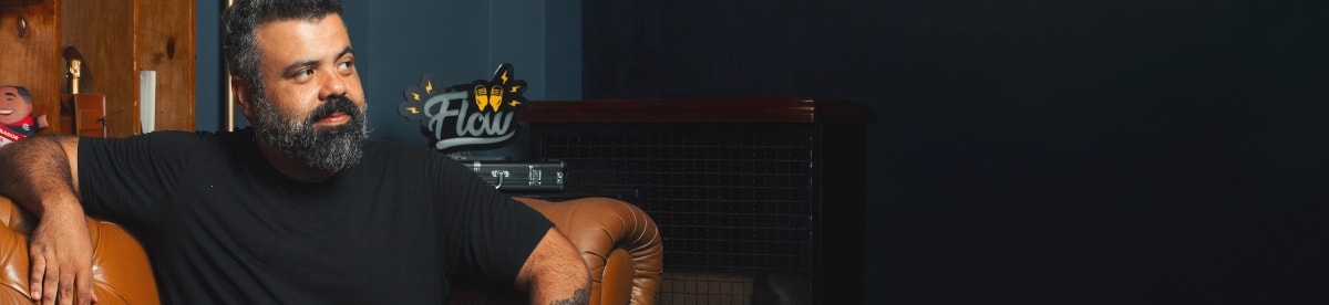 Igor 3k, apresentador do Flow Podcast, com barba e vestindo uma camiseta preta básica, está sentado em um sofá de couro marrom. Ele olha para o lado direito e, ao fundo, vê-se o letreiro luminoso com o logo do Flow. Imagem da campanha Manual de Sobrevivência do Guarda-Roupa Masculino da OQVestir.