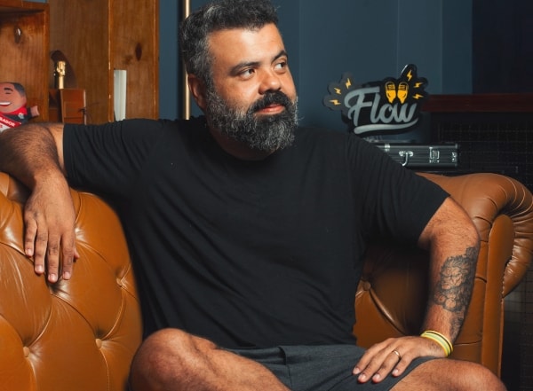 Igor 3k veste OQVestir - Parceria Flow Podcast e Manual de Sobrevivência do Guarda-Roupa Masculino Fotografia de Igor 3k sentado em um sofá de couro marrom clássico, usando uma camiseta preta e bermuda escura. Ele está com o braço esquerdo apoiado no encosto do sofá, exibindo uma tatuagem, e o logo do Flow Podcast aparece desfocado ao fundo. Imagem para a campanha de moda masculina da OQVestir.