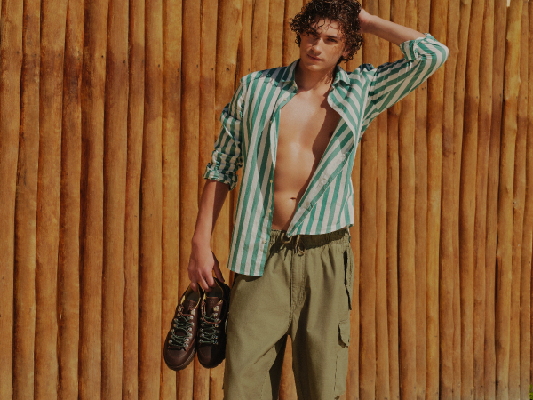 Look masculino casual de verão com camisa leve de listras verticais, calça cargo verde e calçado de couro marrom em cenário com painel de bambu.